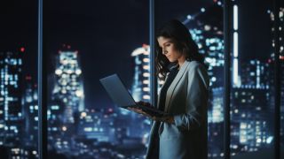 Woman standing in a room at night time with a backdrop of a city, while holding a laptop and using it with one hand 