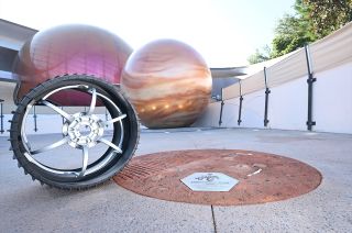 a black rover wheel stands on concrete, in front of a model of the planet jupiter.