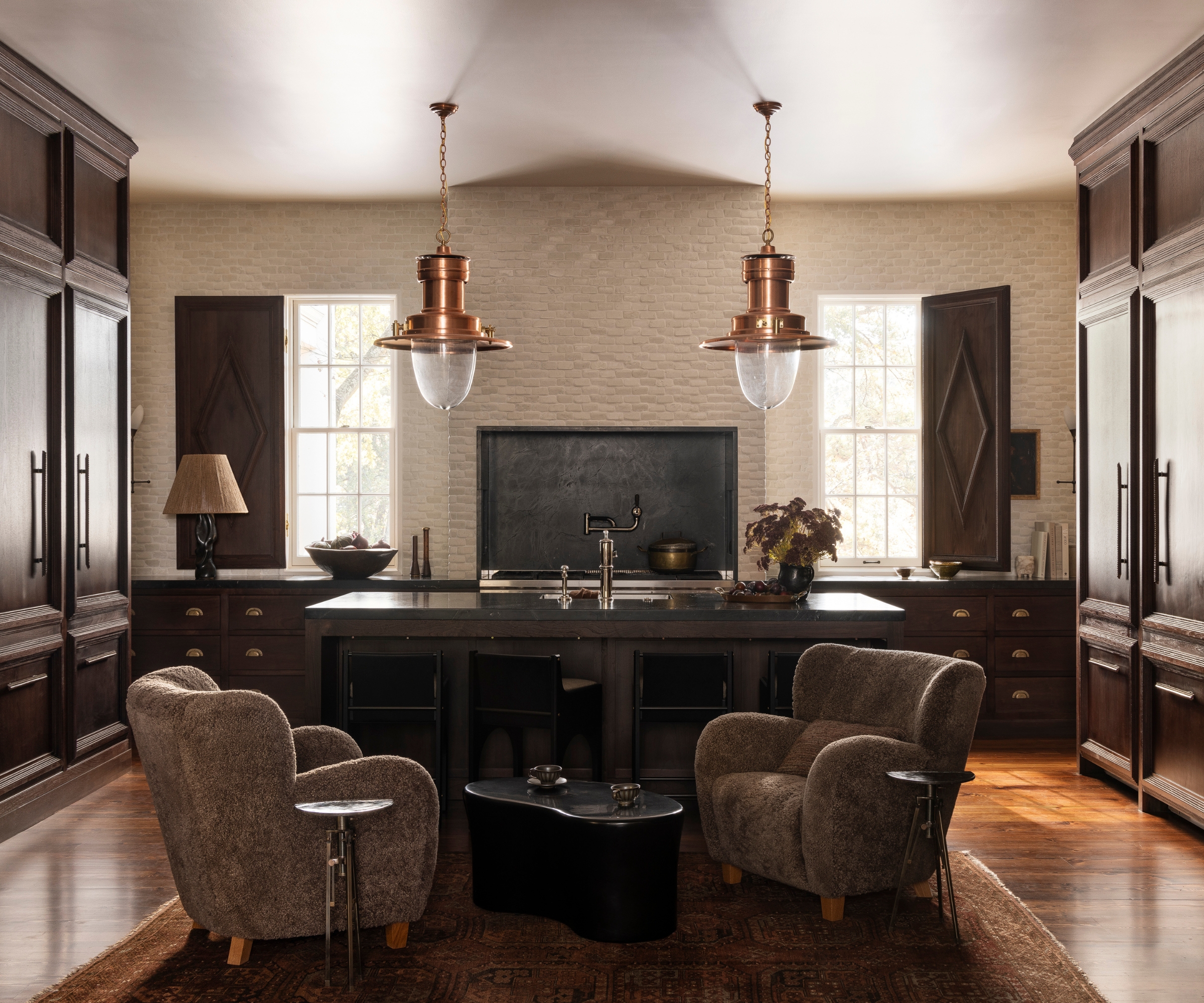A moody kitchen with dark wood cabinets, black countertops, brass pendant lights, low-back counter stools, and a mini seating area with two armchairs and a coffee table