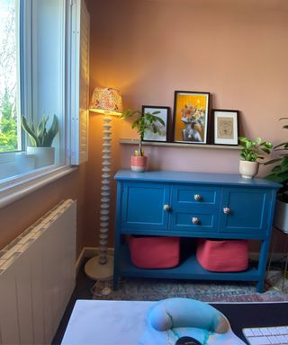 Terracotta small home office with blue sideboard, red storage baskets underneath, a white Bobby lamp, green small plants and pictures on a wall-mounted shelf