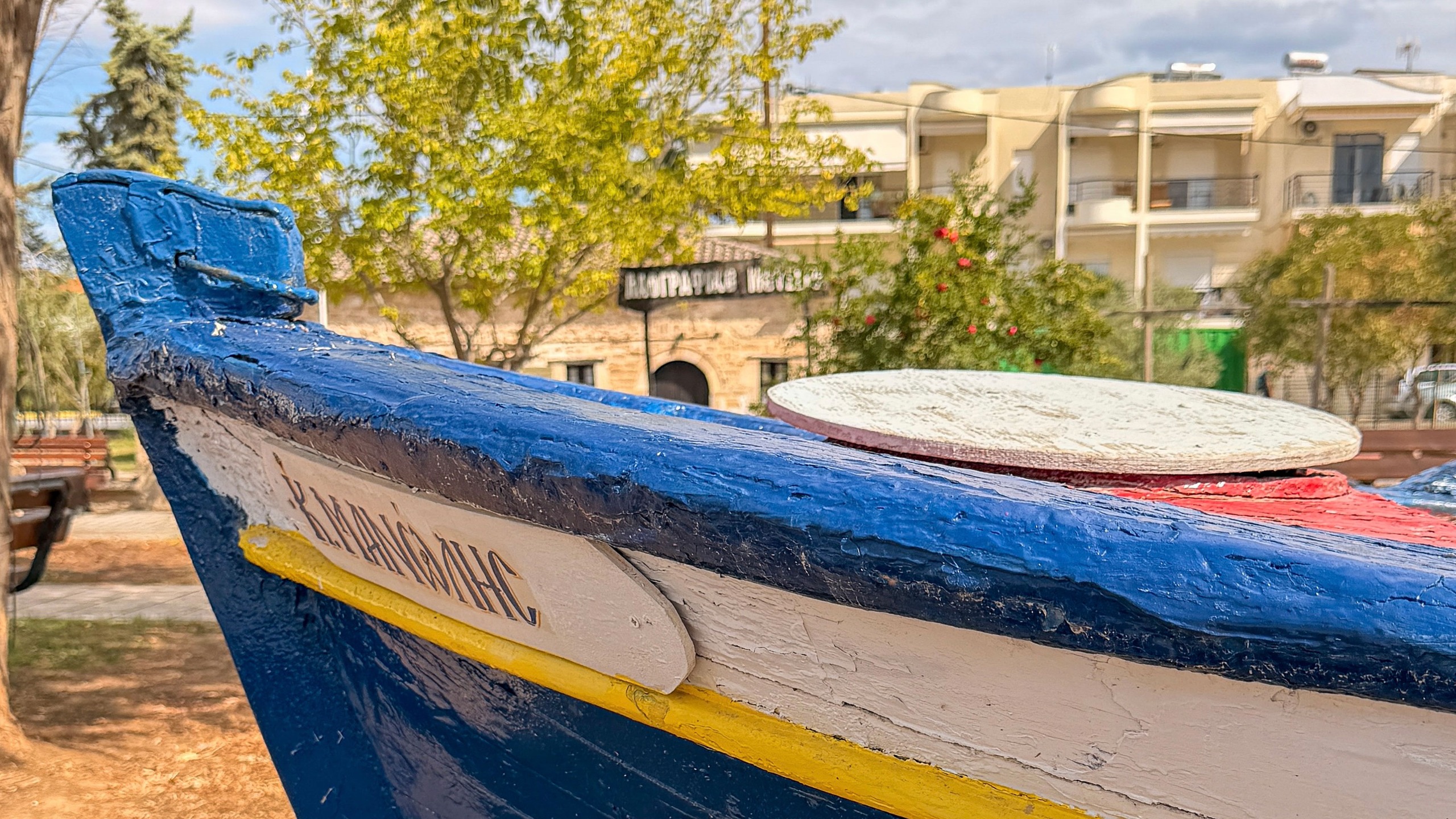 Painted wooden boat with Greek writing on land in front of buildings and trees
