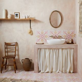 coastal bathroom with pale pink walls, pink and yellow patterned splashback and floor tiles, curtain under sink, vintage mirror, vintage chair, rug, shelf