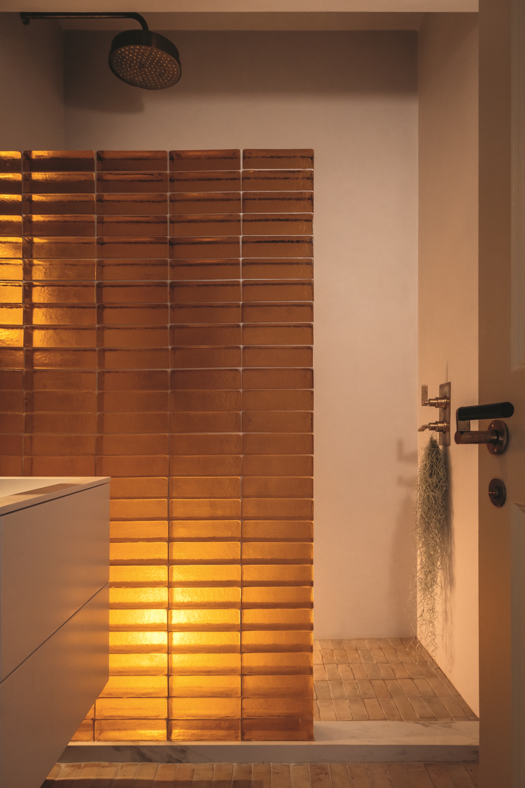 modern bathroom with plaster pink walls, brick floors, amber glass brick wall, overhead shower, and plant hanging from taps