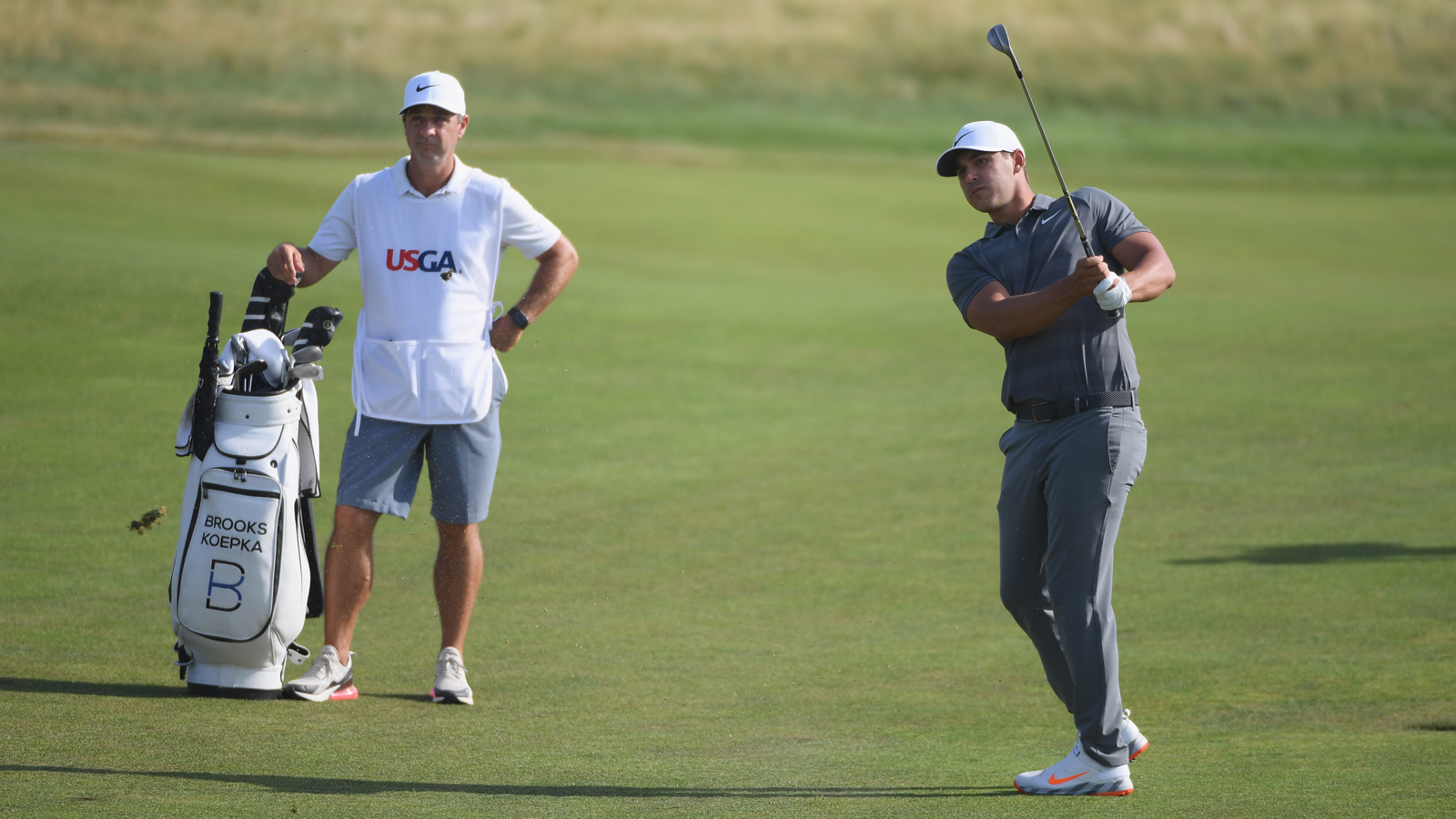 Brooks Koepka plays an approach shot as caddie Richard Elliott looks on during the final round of the 2018 U.S. Open at Shinnecock Hills Golf Club