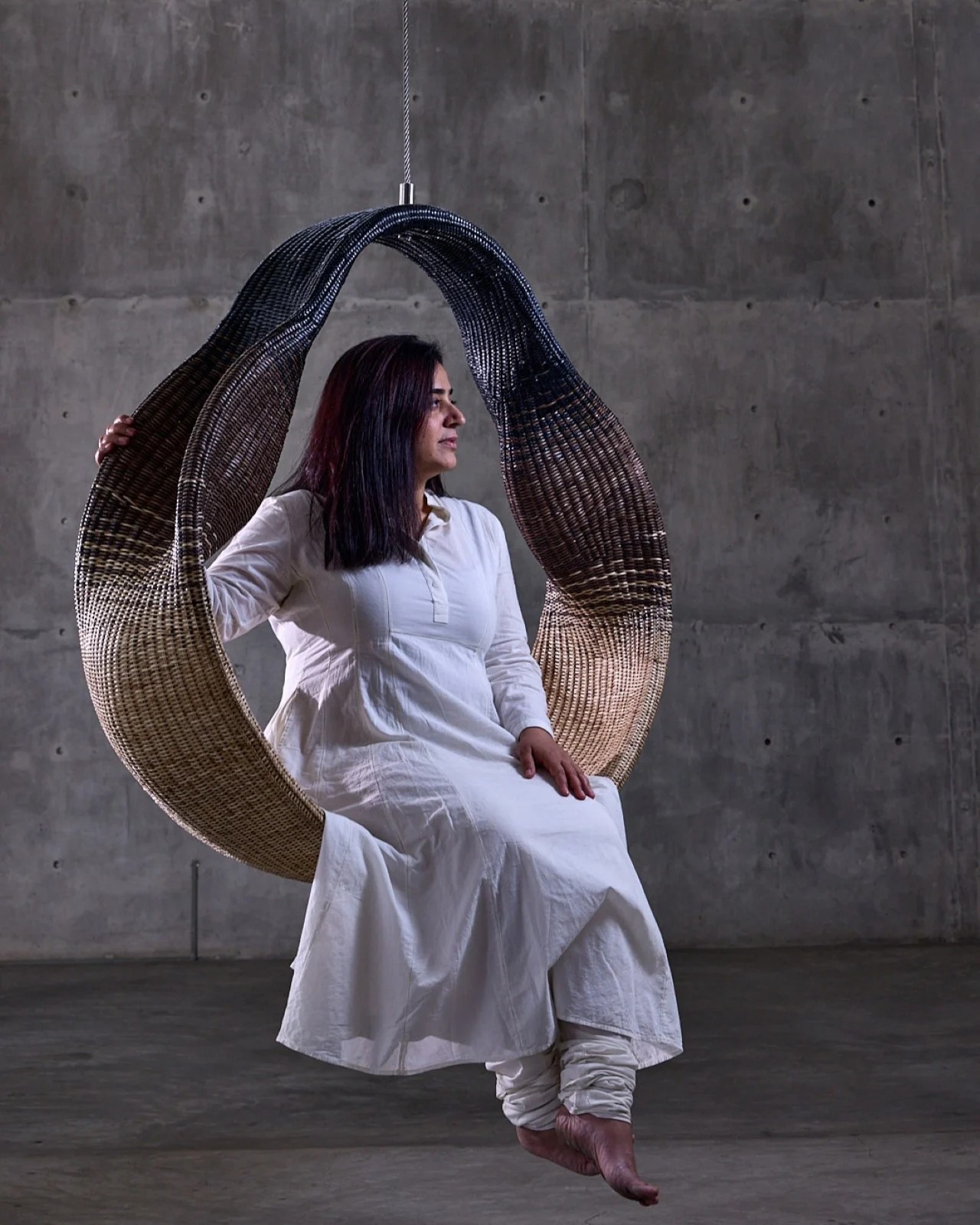 Woman in full-white, loose linen outfit, sitting in an oval-shaped wicker creations floating from the ceiling in a cement room, captured sideways.
