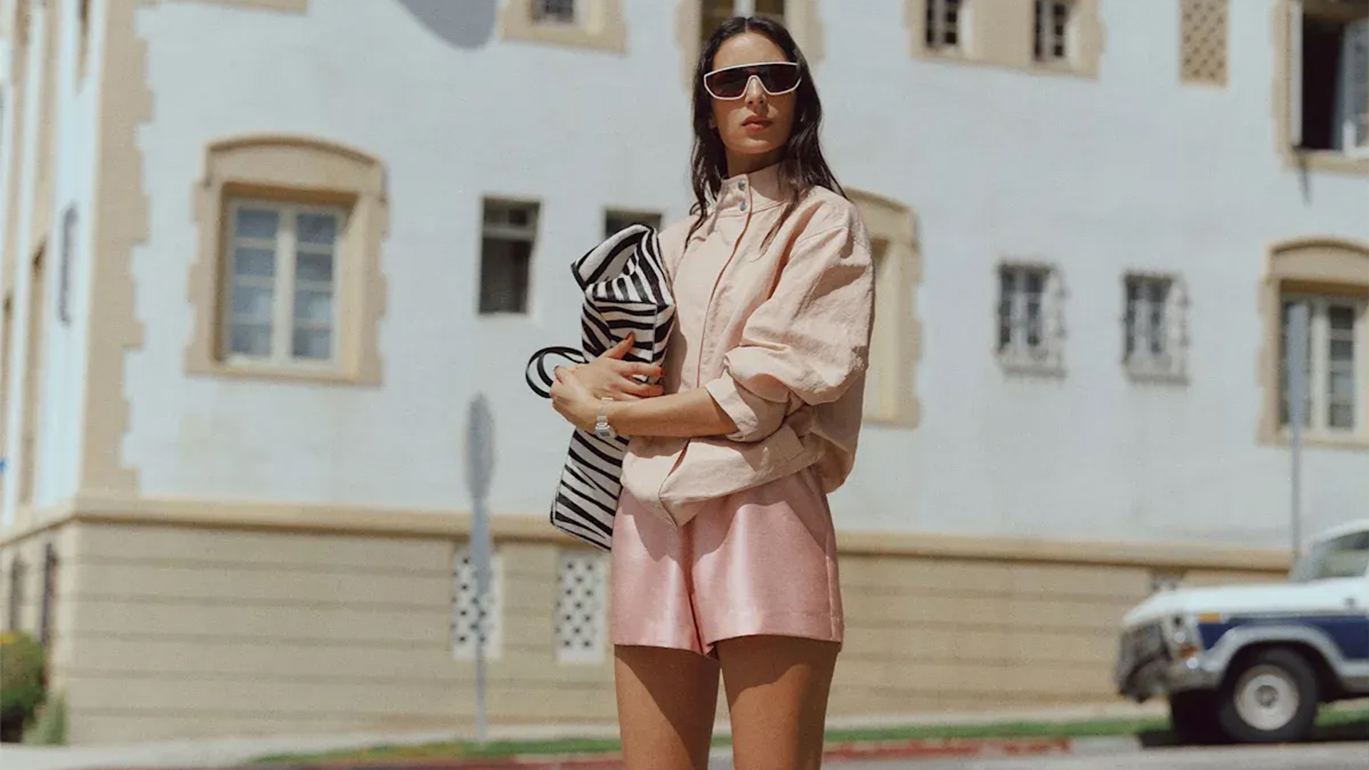 Model wears funnel neck top, satin pink shorts, white wedge heels, and zebra print tote while posing for the camera. 