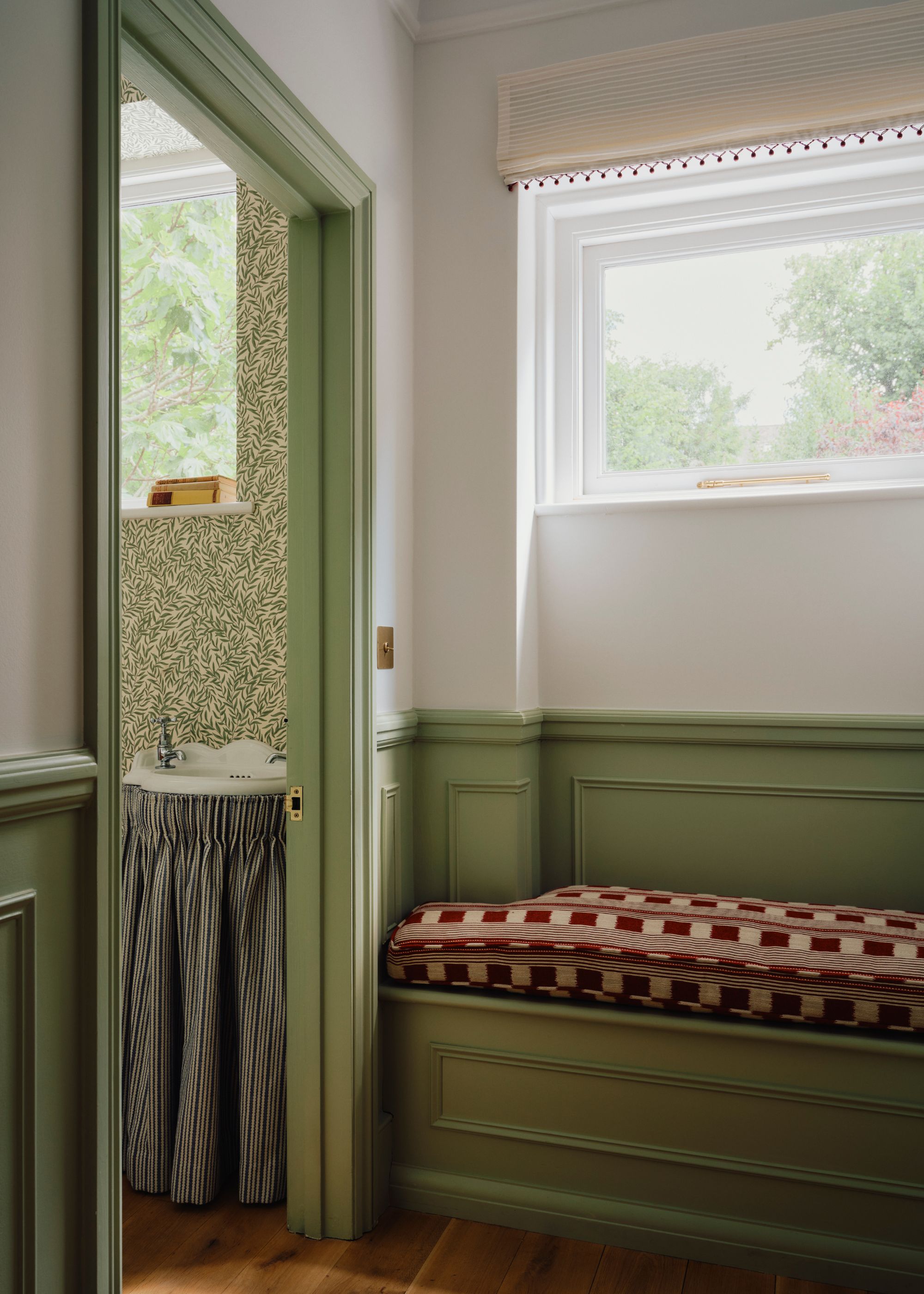 A hallway with traditional panelling painted a mint green color, with a matching doorframe leading into a green wallpapered powder room. White paint above the panelling, with a window and a bench seat with a red patterned cushion.