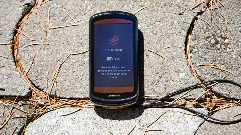 A black Garmin edge 1040 solar sits on a paved floor. The solar status page shows on the screen, showing the sunlight intensity with advice on how to charge it faster