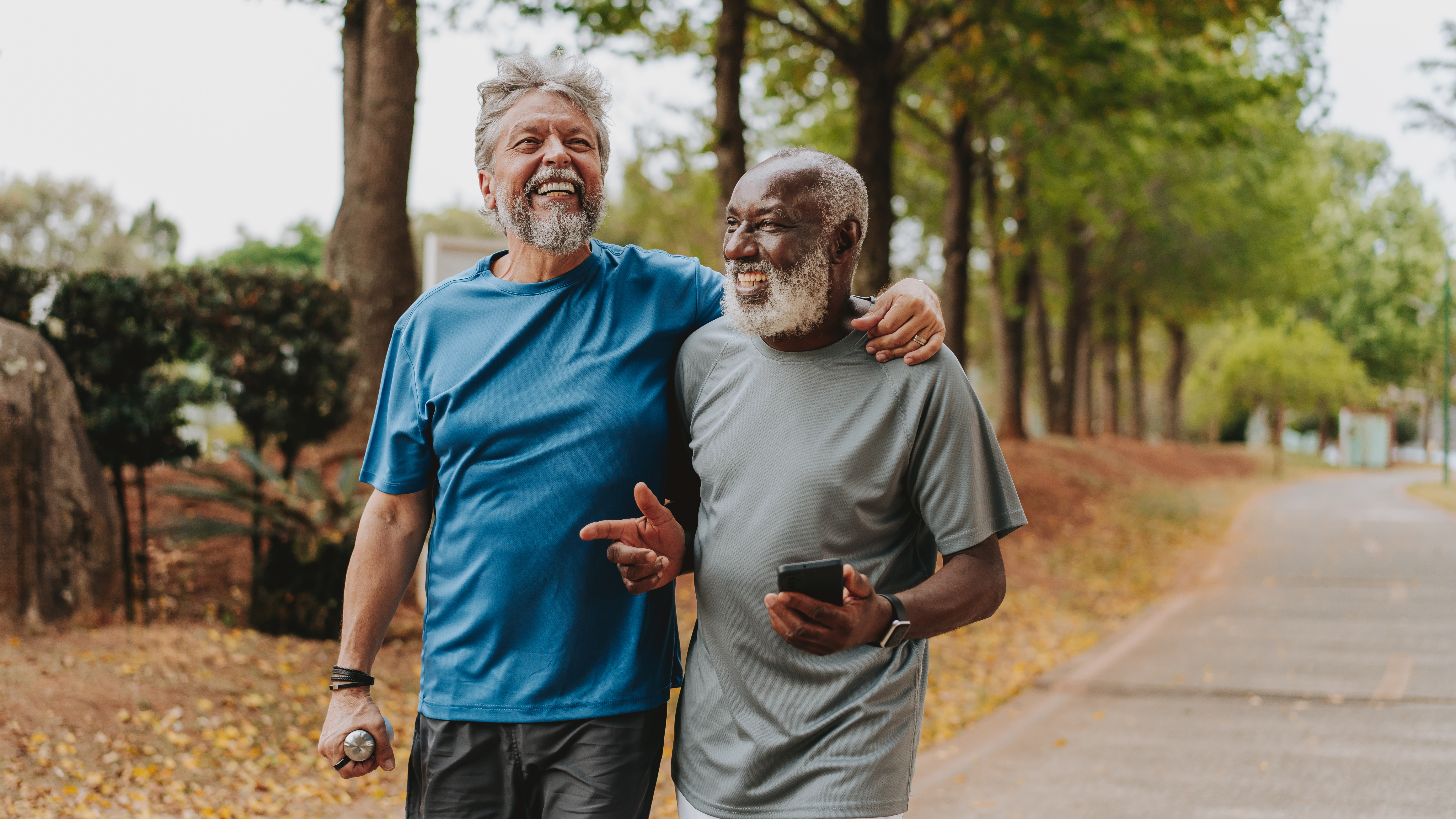 Two men walk next to each other, laughing. One of them has thrown his arm around the other. Behind them we see rows of trees and brown leaves on the ground.