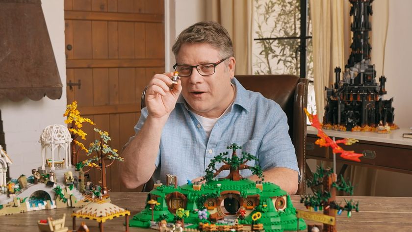 Sean Astin holds up a Samwise Gamgee minifigure while seated at a table in front of Lego The Shire, with other Lord of the Rings kits around him in a cottage room