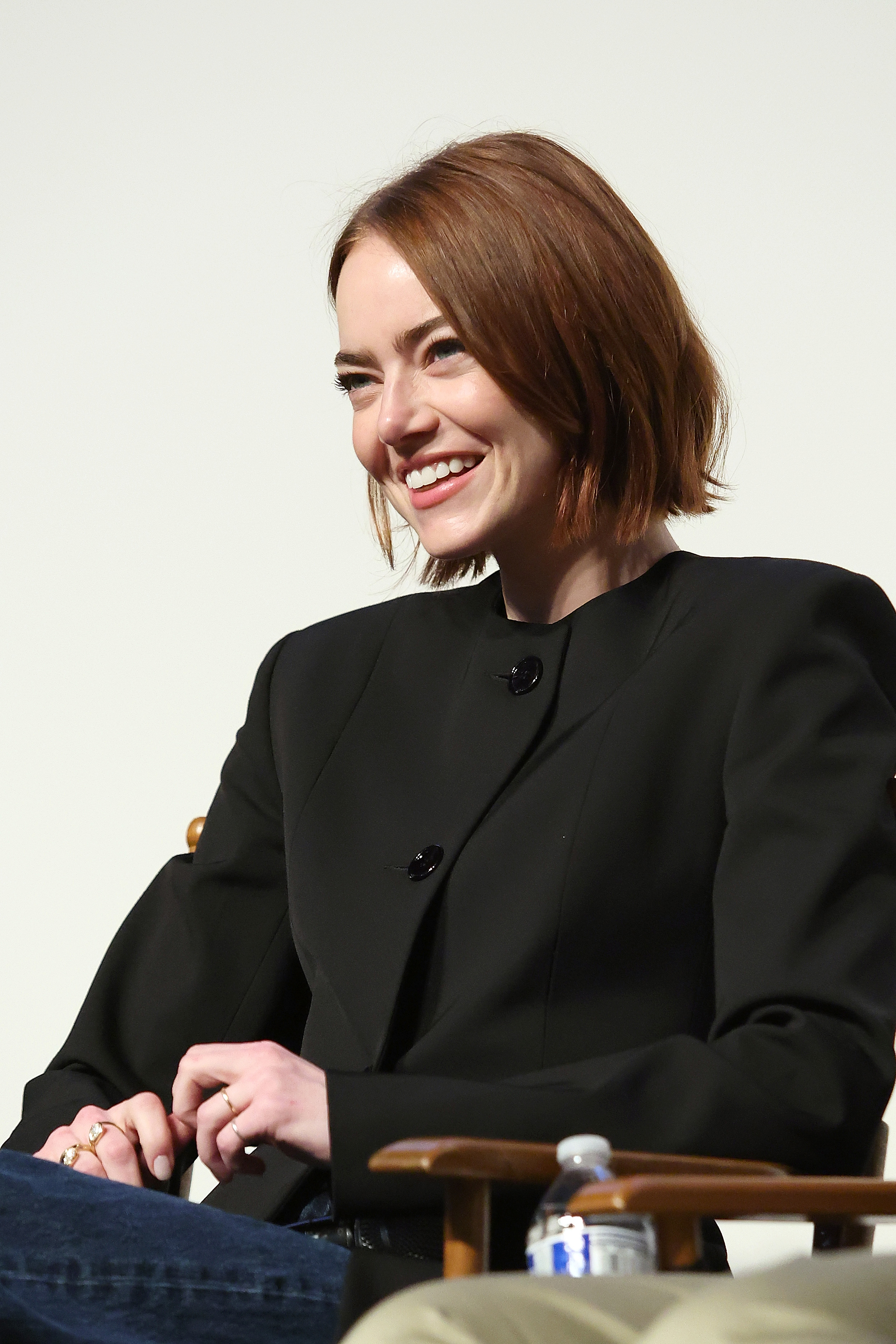 a white woman with short, red hair wearing a black blazer and blue jeans. she is sitting in the photo