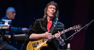 Steve Hackett is feeling his solo as he performs live with his gold-top Fernandes Les Paul copy. He wears a black jacket and a red T-shirt.