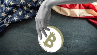 A hand holds a bitcoin over the US flag draped over a slate board.