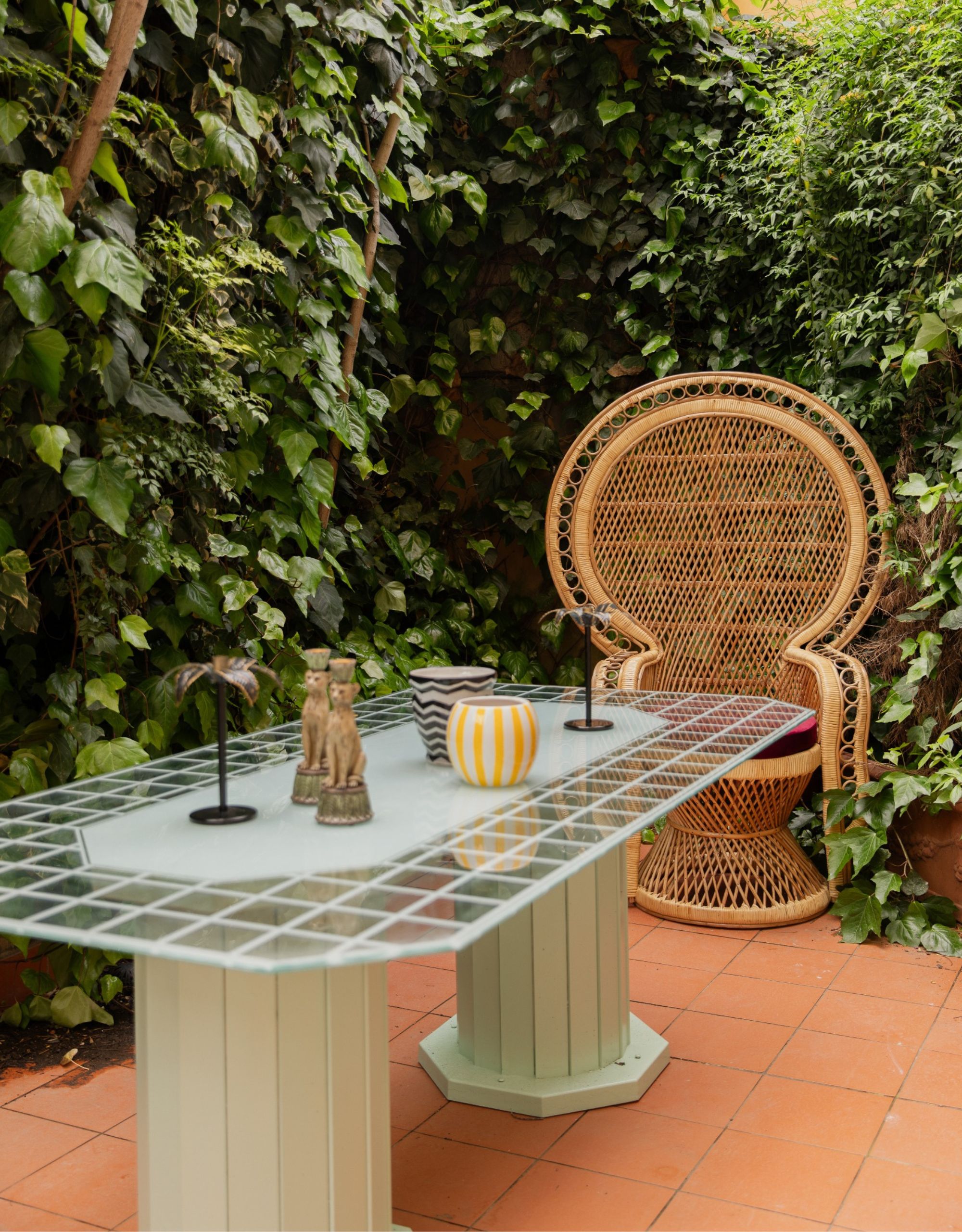 A small outdoor area with terracotta flooring, a glass table with decor on top and a round wicker peacock chair surrounded by foliage