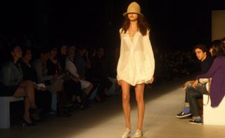 Model on runway wearing a partially-sheer shirt and low hat