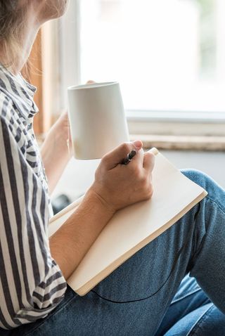 A woman sits with a mug of coffee, a pen and a blank journal page