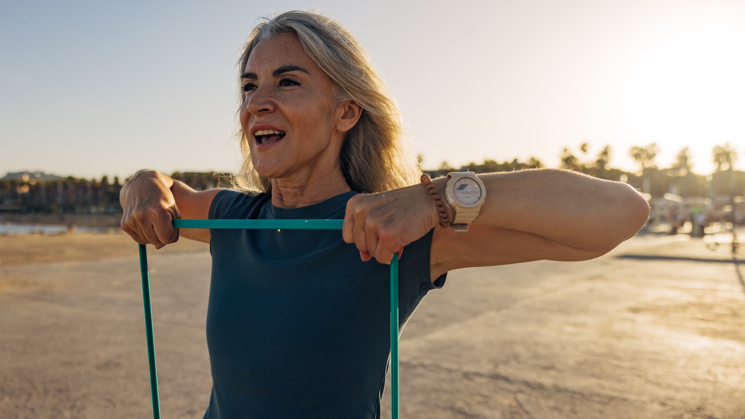 A woman exercises outside, holding a resistance band up to her shoulders and holding it taut in her hands.