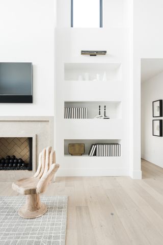 Living room with pale wood floor, white walls and built in shelving, fireplace with TV above, wood chair carved in shape of hand and neutral patterned rug