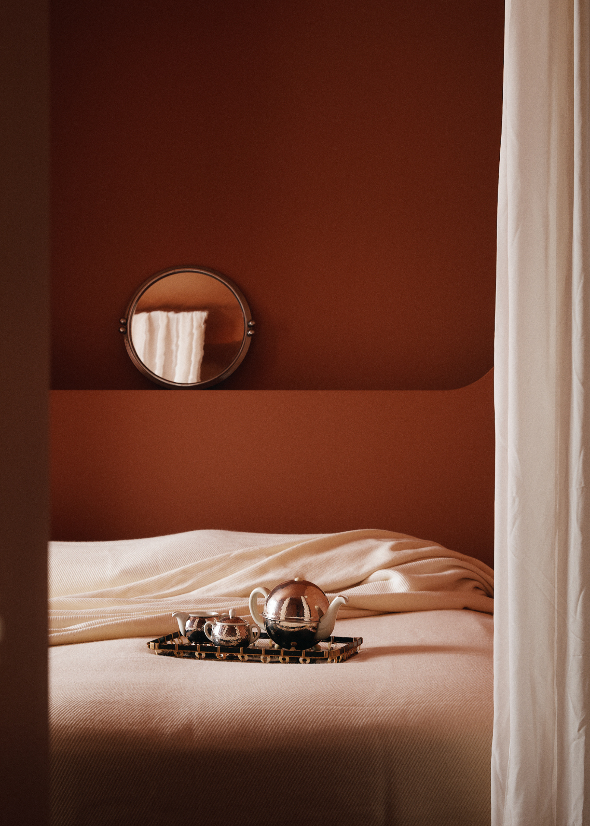A dusty pink bedroom with a circular mirror above the bed, a chrome tea set on a tray on the bed, and soft pink sheets
