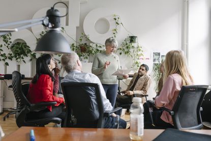 A woman holding a sheet of paper and talking while surround by her colleagues sitting in chairs at the office together