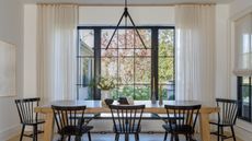 Dining room with large window and black crittal design. There is a large eight seat dining table with black spindle back chairs, floor to ceiling sheer curtains and landscaped yard in view with red maple tree