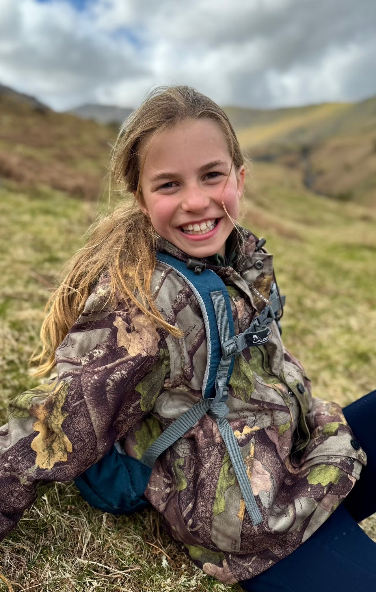 Princess Charlotte wearing a camo jacket sitting on a hill and smiling on her 10th birthday