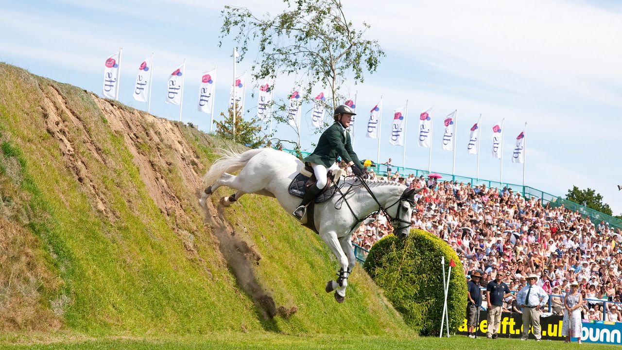A grey horse jumps off the front of the Hickstead Derby bank with a packed grandstand of fans looking on