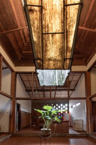 Interior image of Demas Nwoko house in Nigeria, high wooden beam ceiling, glass centre piece letting in light, wooden framed room, wooden floor, potted rubber plant, perforated wall letting in light, narrow horizontal windows, doorways, counter top table and tall stools