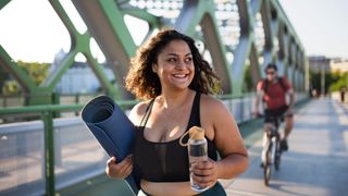 Woman walking holding exercise mat and water bottle with male cyclist in the background.