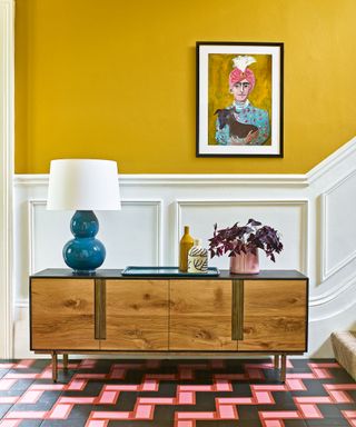 Hallway with yellow painted walls, white painted paneling, wooden sideboard with flowers, table lamp with white shade, striking pink and black
