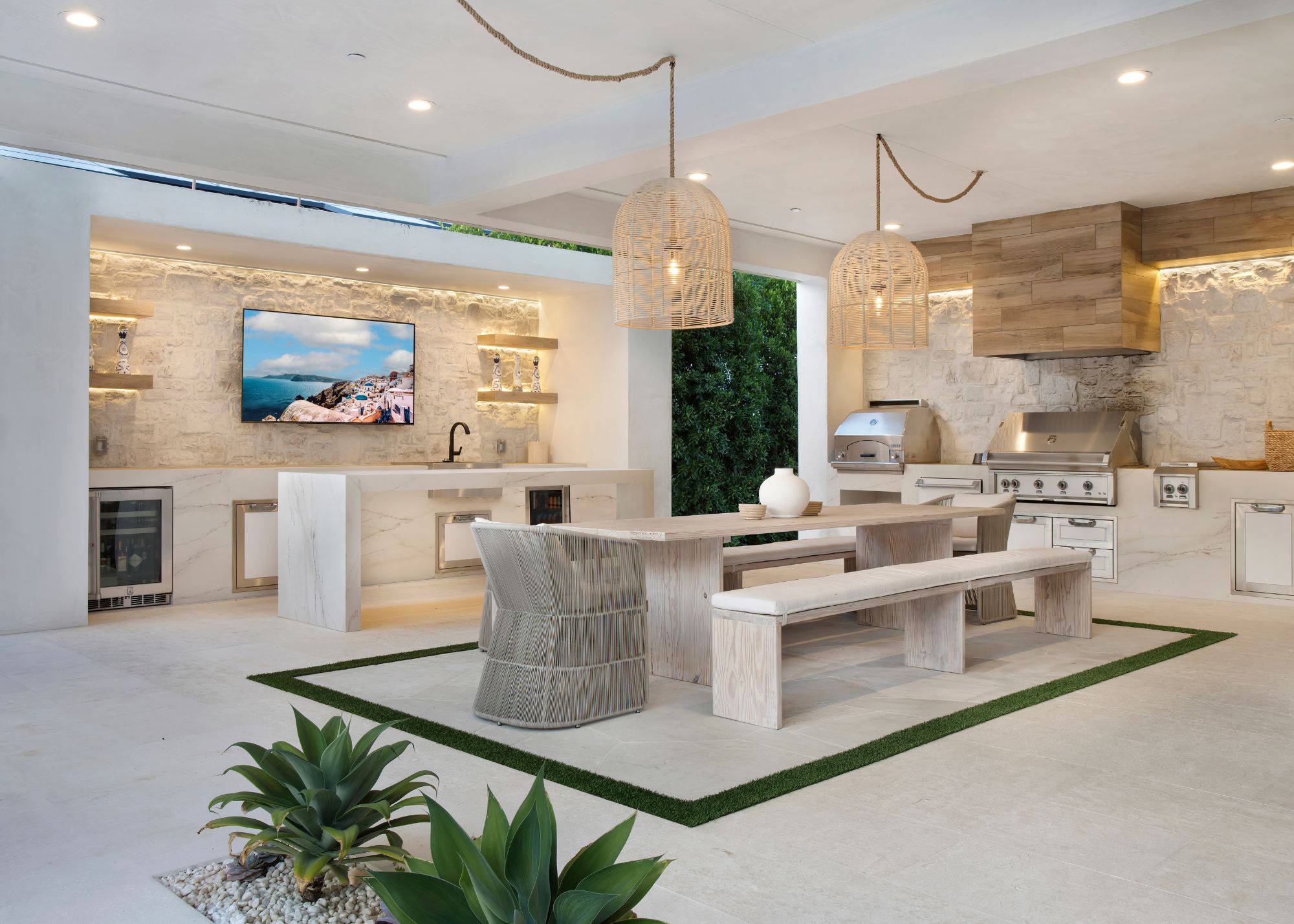 A luxurious outdoor kitchen with two countertop areas and a tv, with a bench seating area in the center with a tiled rug border around it and two rattan pendant lights hung with rope from the ceiling