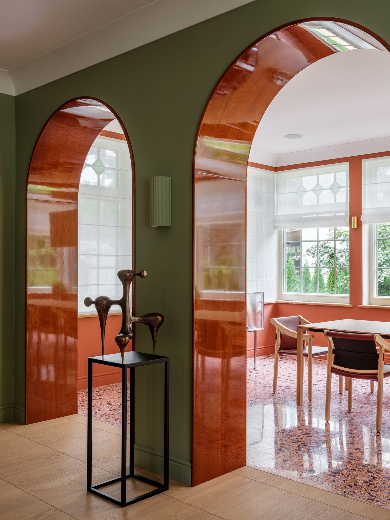 Image of a green living room with arched doorways that lead into an orange-themed dining room. The floors of the dining room are orange terrazzo. There are black and wood dining chairs around the dining table and a white paper lantern overhead.