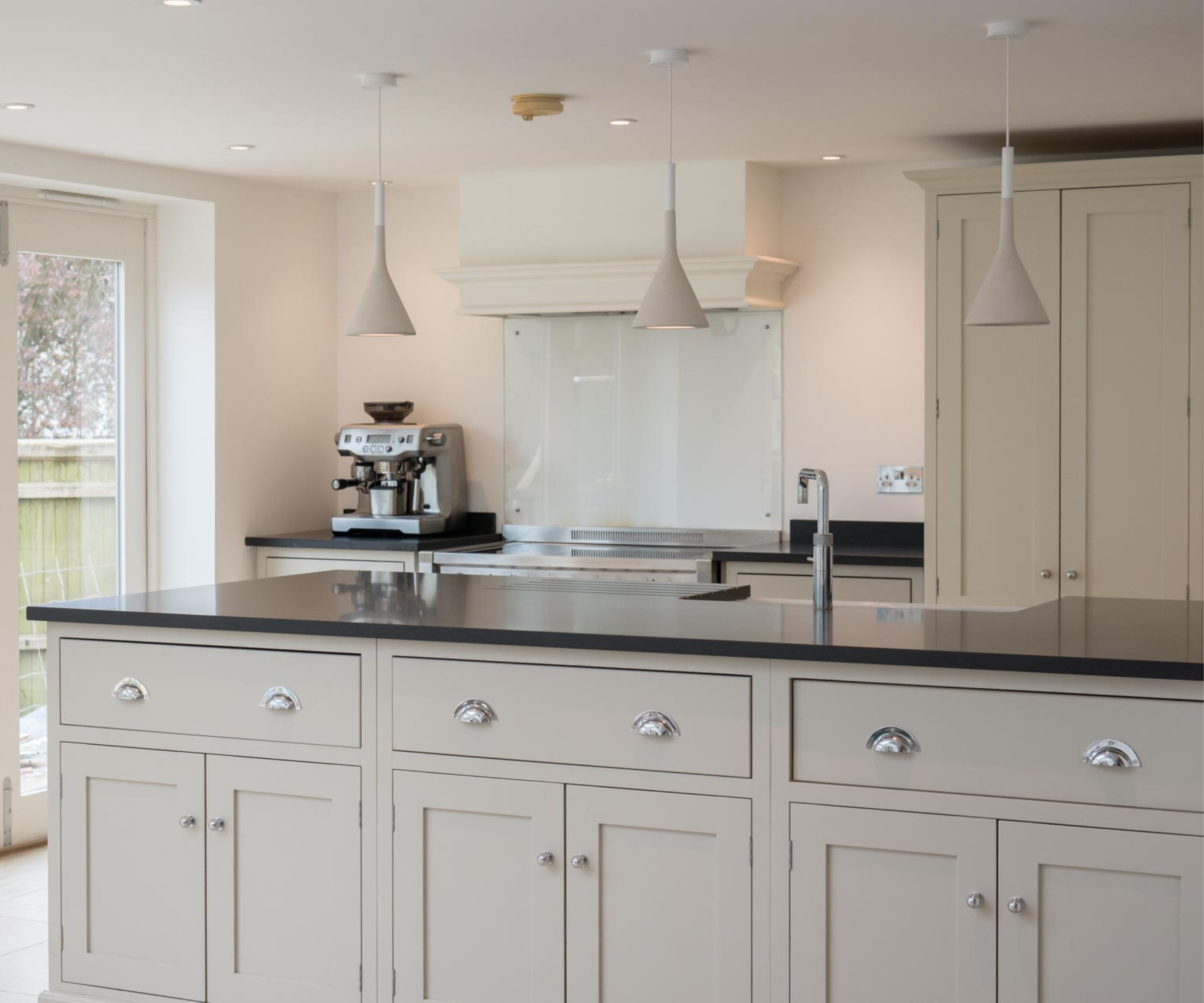 A clean, modern kitchen with light grey cabinetry and a large island topped with a dark work surface. Slim pendant lights hang overhead. Glass doors open to a garden, filling the space with natural light and a calm, minimalist feel.