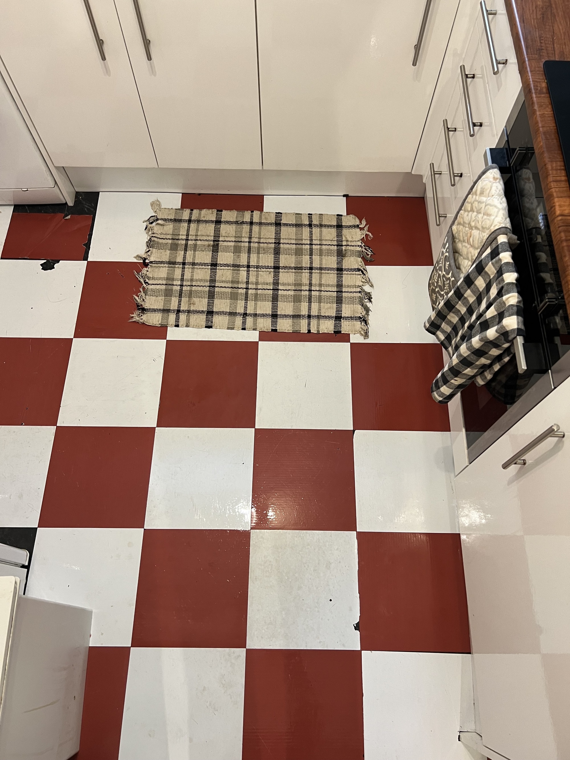 Image of a kitchen floor that has red and white peel-and-stick tiles. Some of them are coming up. There is a green, beige, and black plaid rug.