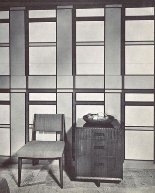 A black and white photo of a chair next to a chest of drawers in front of a wall with a pattern made of squares and rectangles on it.