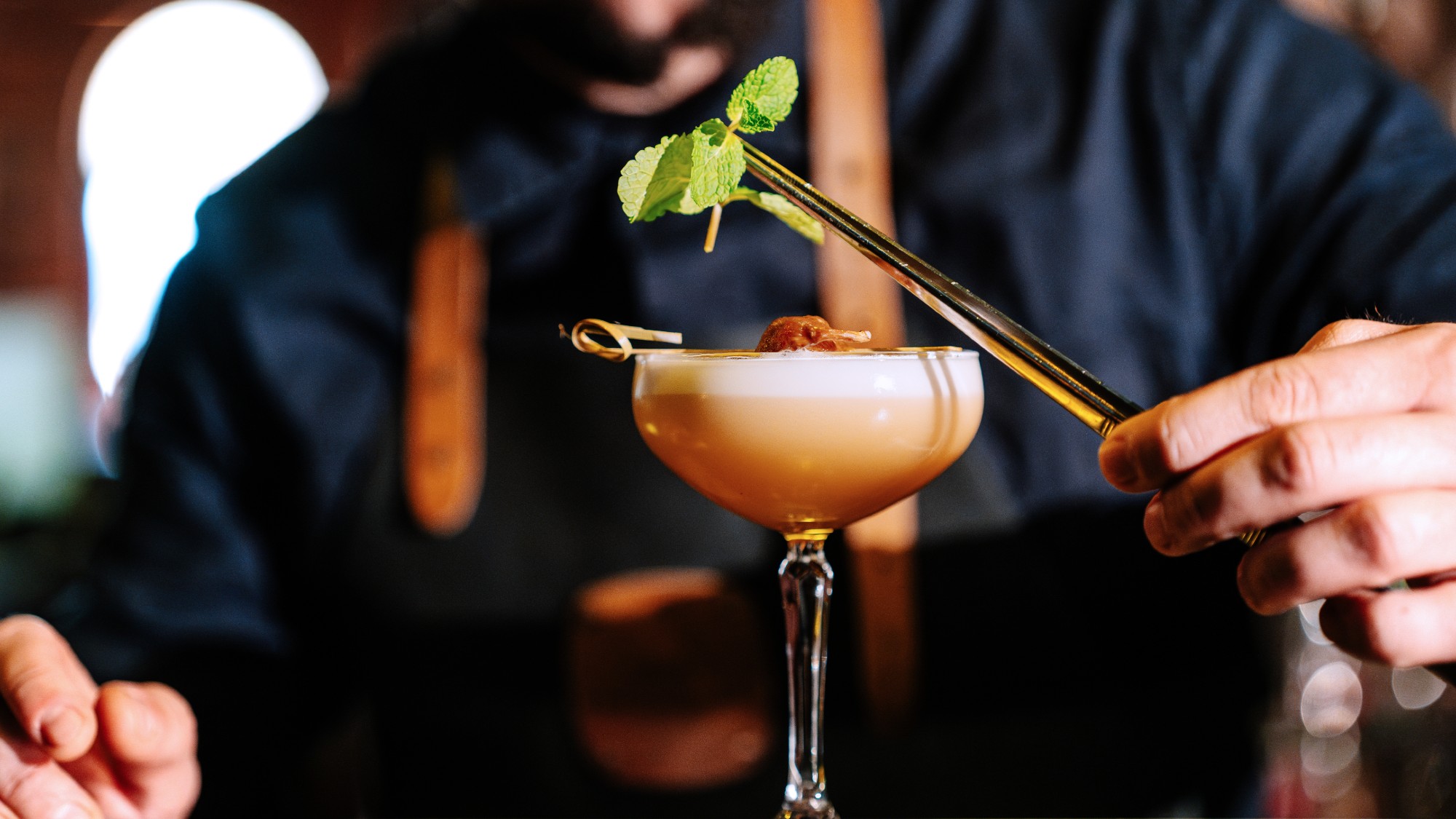direct shot of a bartender in a dark blue shirt and leather overalls garnishing a frothy orange cocktail with a mint sprig. he is using gold tweezers.