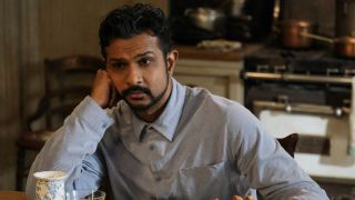 Jay leaning on his left hand while sitting at a table in Ghosts.