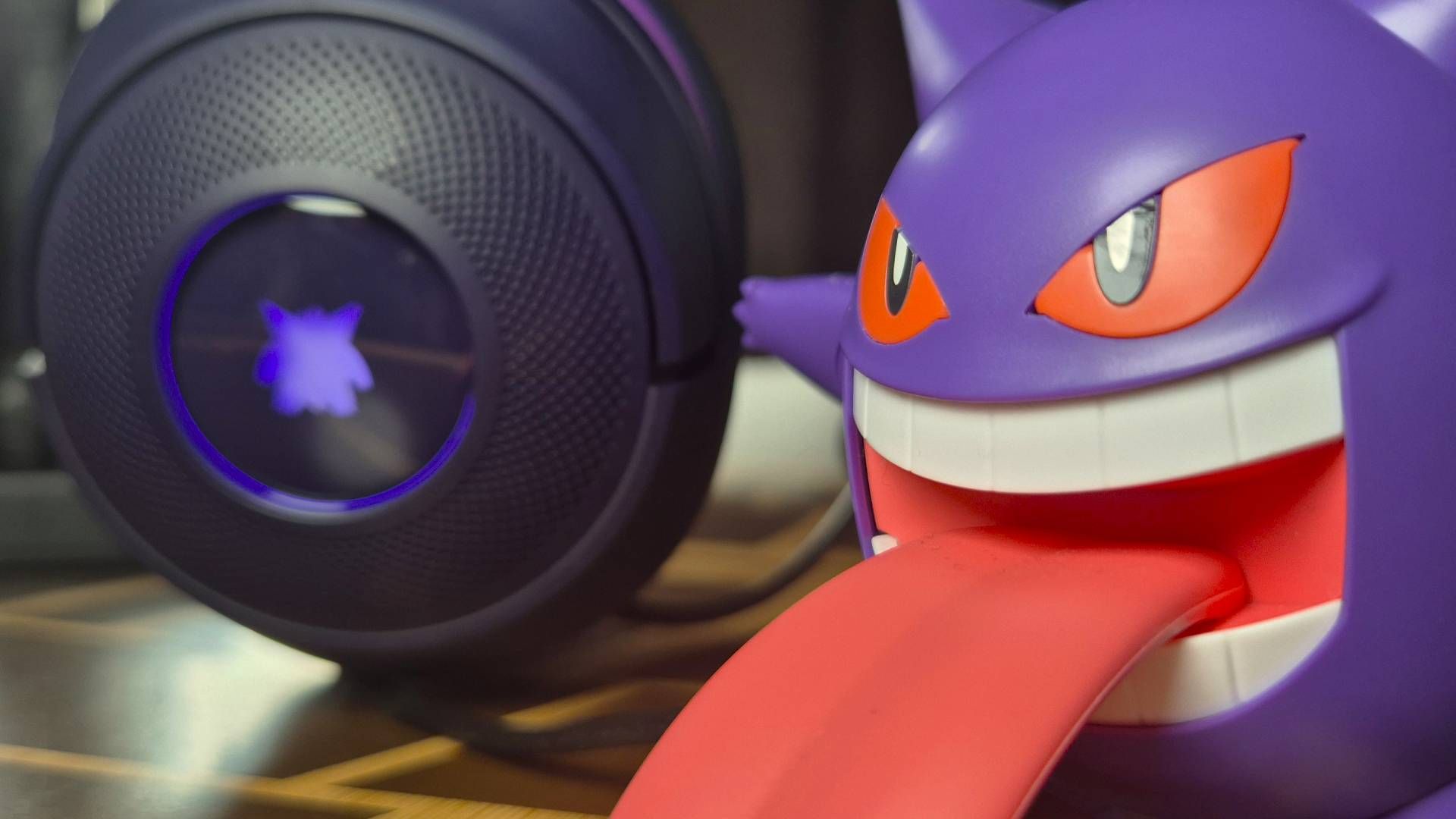 Photo of a close up of a toy of the Pokemon Gengar to the right of the frame, with a purple headset with a glowing Gengar silhouette in the background.