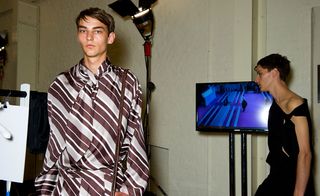 Two guys wearing the JW Anderson S/S 2015 collection. On the left, the guy is wearing a brown and white striped garment with a scarf/tie as part of the garment. Next to him a guy walking past is wearing an off the shoulder black garment