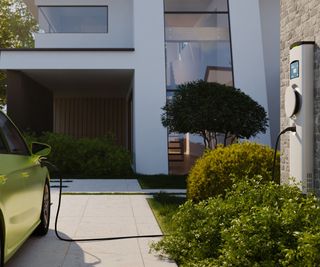 An electric car charging point mounted to a wall at a home charging a green car on a driveway. A modern home in the background