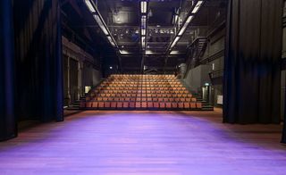Interior image of the Cultural Centre, Dordrecht, wooden stage floor, rows of auditorium seating, high dark ceiling with framework of stage lights, ceiling lighting, steps at either side of the seats, grey stone walls, dark coloured stage curtains