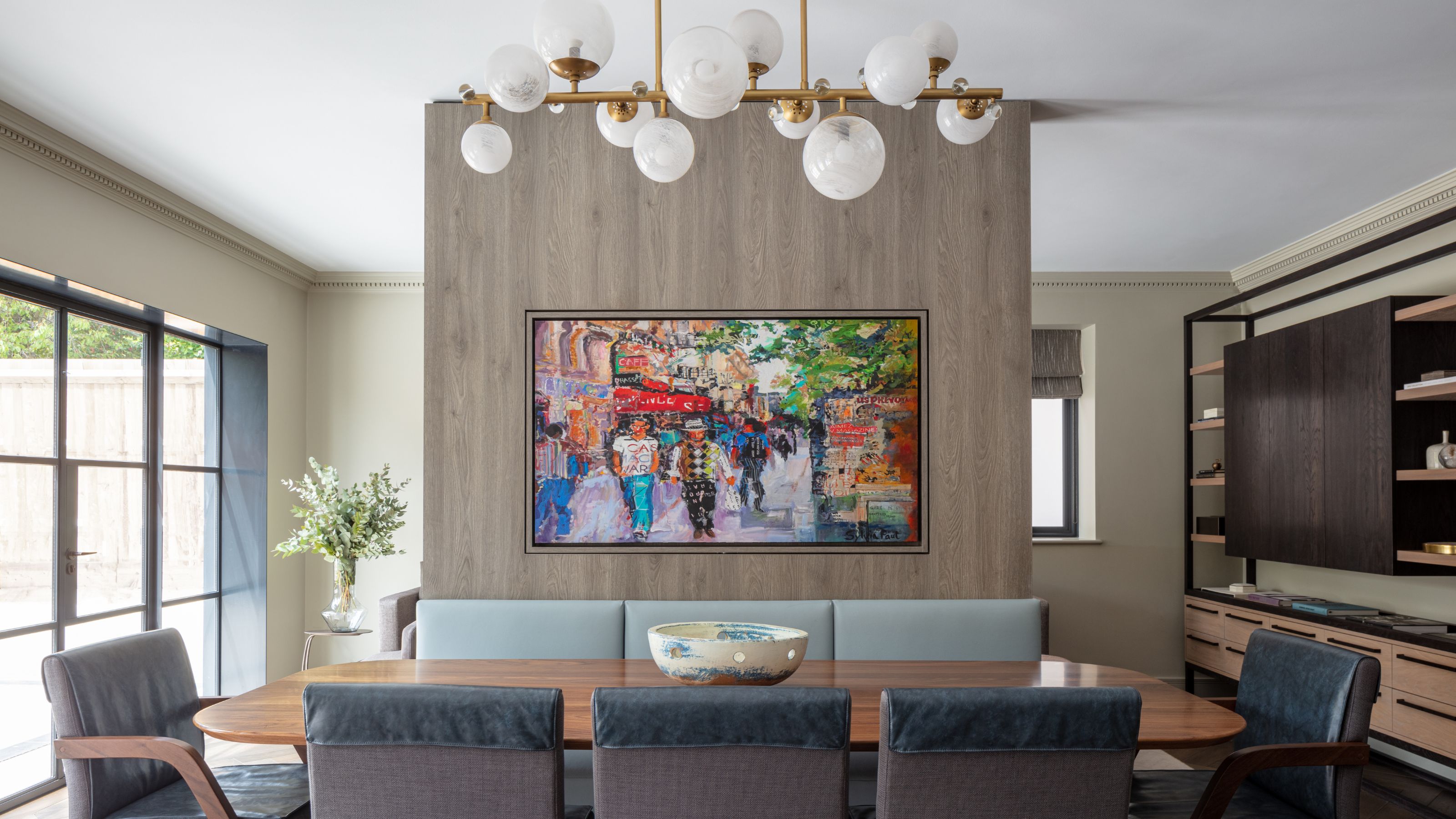 A dining area with large anthracite windows and doors as well as a view of a large artwork placed in the middle of the room. Above the dining table is a large pendant light with gold hardware
