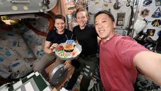 a man in a red shirt takes a selfie with two women in dark colored shirts as they hold up a makeshift cake with a "200 Days" embroidered patch mounted atop it.