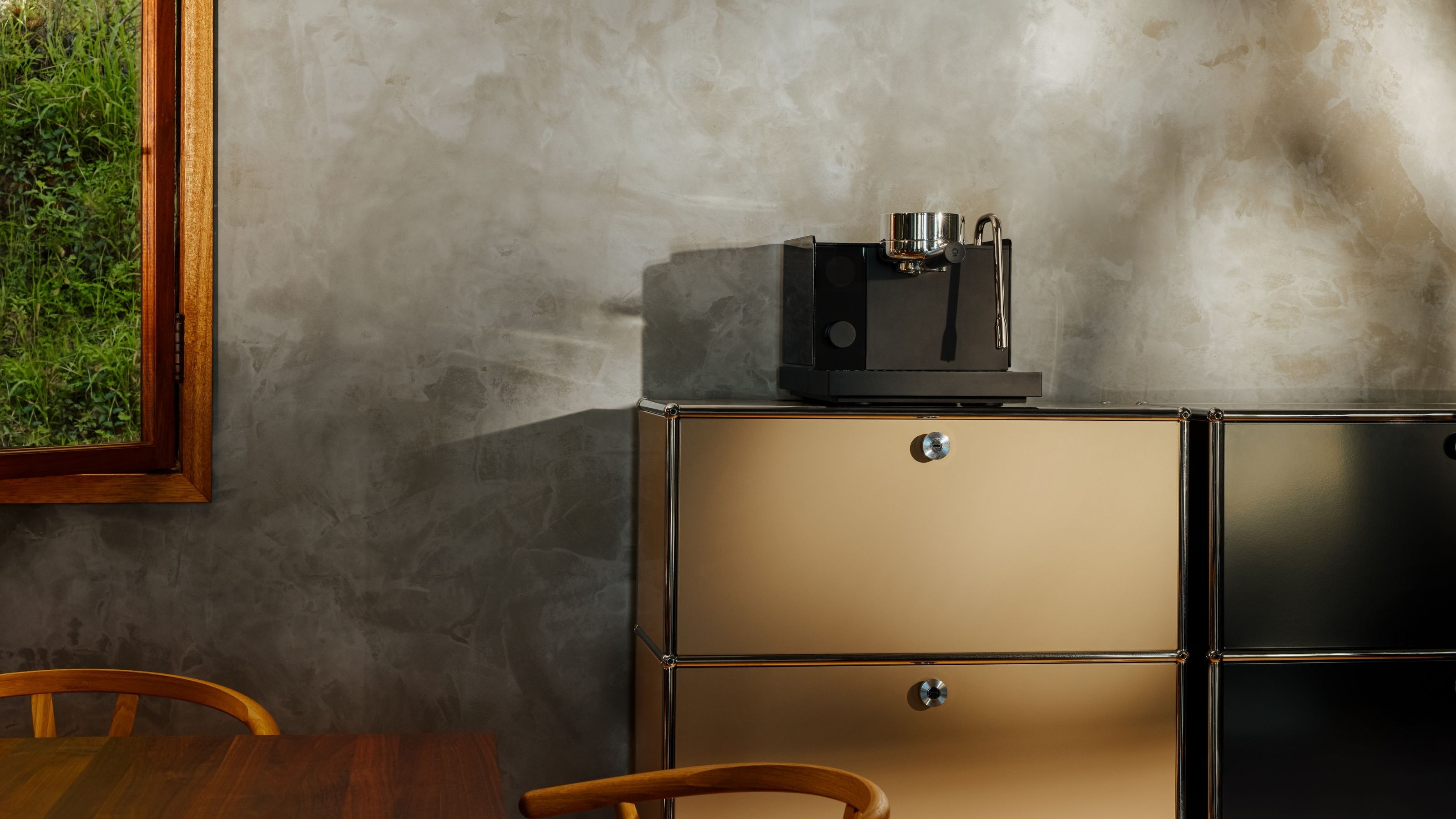 A black Fellow Series 1 espresso machine on a tan, metal cabinet of drawers in front of a gray limewash wall.