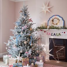 Pale pink living room with frosted faux tree with pastel pink and blue ribbons, neutral baubles, star on wall and in fireplace, candles, falala garland, presents under tree