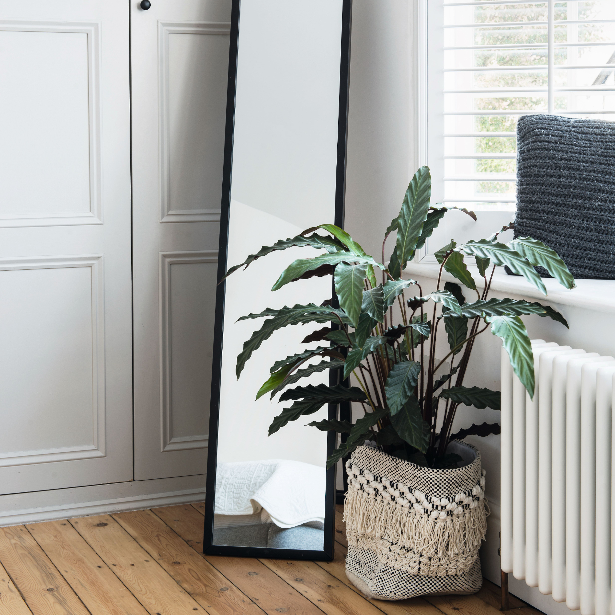 Standing mirror on a wooden floor beside pale grey built in wardrobe and window with venetian blinds