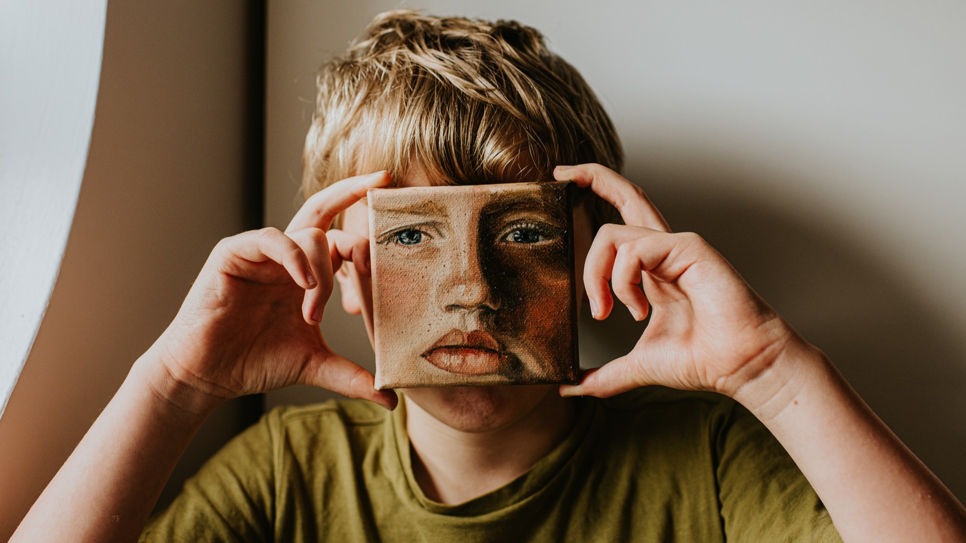 A young lad covers his face with a small oil painting of himself. The expression on the painting is serious.