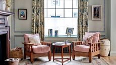 Two wooden arm chairs with pink cushions, either side of a small round wooden table. A large window in the background. 