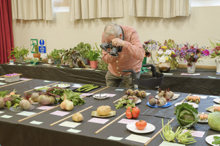 Martin Parr leans over a village hall table laden with prize vegetables and flowers, ring flash mounted on his camera, with his walking frame parked beside him.