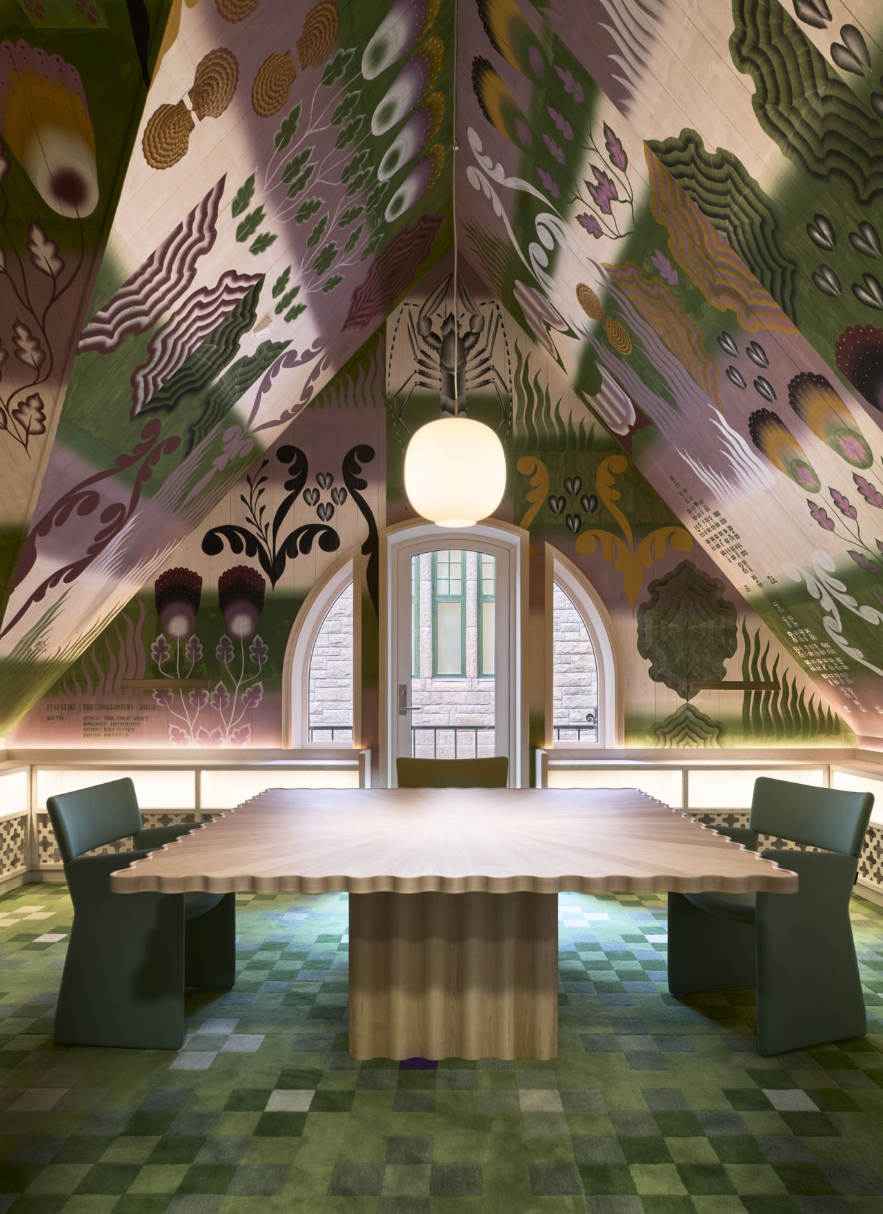 A whimsically decorated, green, gold, and purple-hued space features sculptural minimalist furniture that contrasts with the widely figurative walls of an attic-style room with a pendant light at its center.